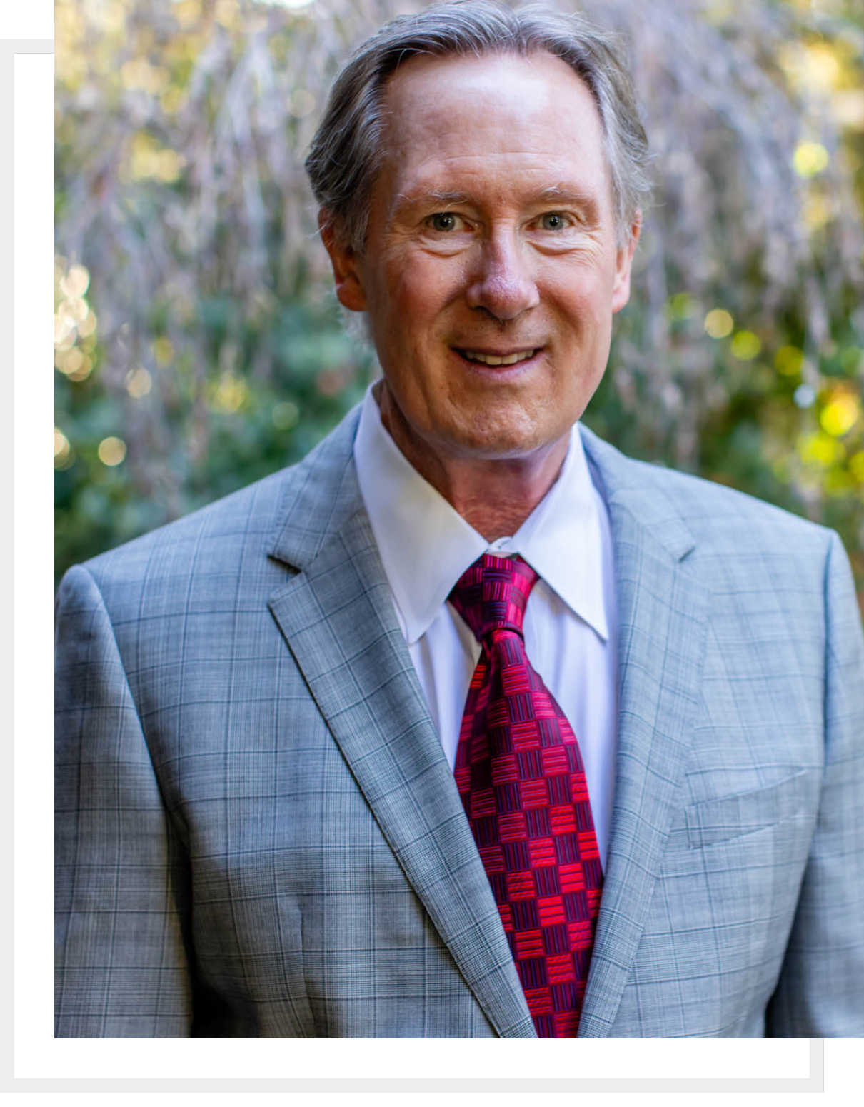 Smiling elderly man in a gray suit and red tie outdoors.