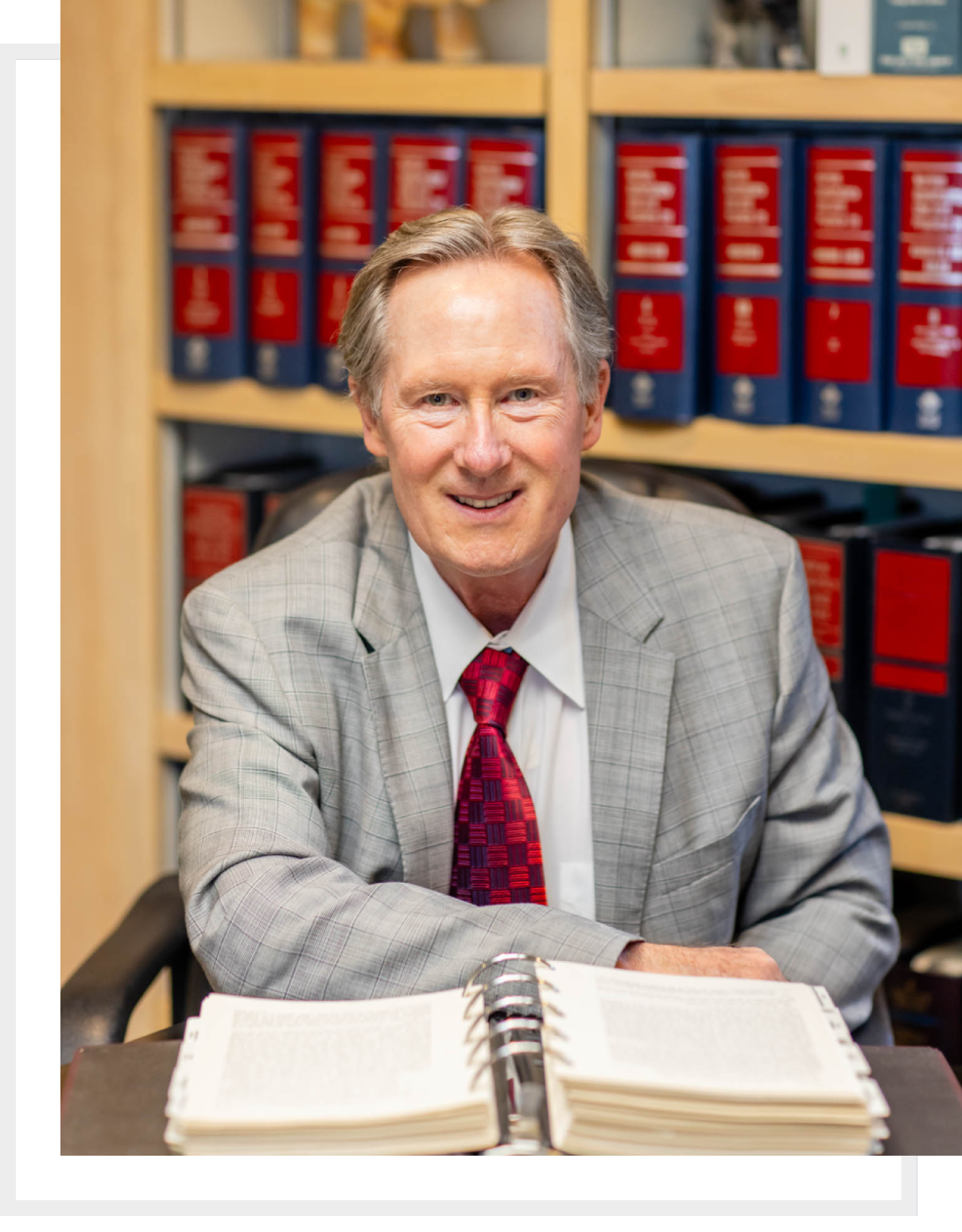 An older man in a suit and red tie sitting at a desk with books.